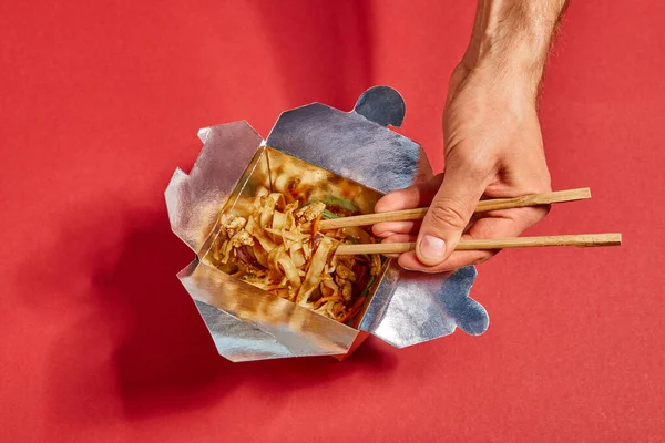 Vista recortada del hombre sosteniendo palillos cerca de fideos picantes en rojo - foto de stock