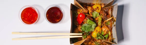 Panoramic shot of sauces and chopsticks near takeaway box with prepared pork and vegetables on white — Stock Photo