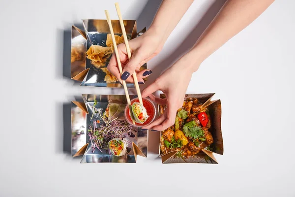 Vista superior de la mujer sosteniendo palillos con dumpling y salsa cerca de cajas de comida para llevar con comida china preparada en blanco - foto de stock