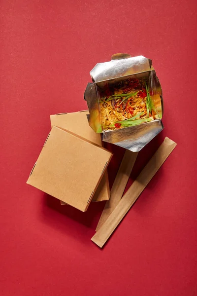 Top view of chopsticks in paper packaging near takeaway carton boxes and tasty noodles on red — Stock Photo