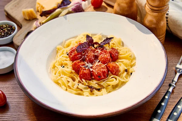 Вкусная паста с помидорами, базиликом и пармезаном подается в круглой тарелке на деревянном столе при солнечном свете — стоковое фото