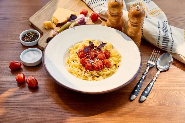 Deliciosa pasta con tomates servidos en mesa de madera con cubiertos, servilleta, condimentos e ingredientes a la luz del sol - foto de stock
