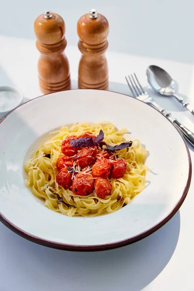 Delicious pasta with tomatoes served with cutlery, salt and pepper mills on white table in sunlight — Stock Photo