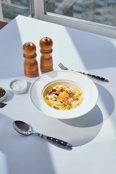 Delicioso macarrão carbonara servido com talheres, moinhos de sal e pimenta na mesa branca à luz do sol — Fotografia de Stock