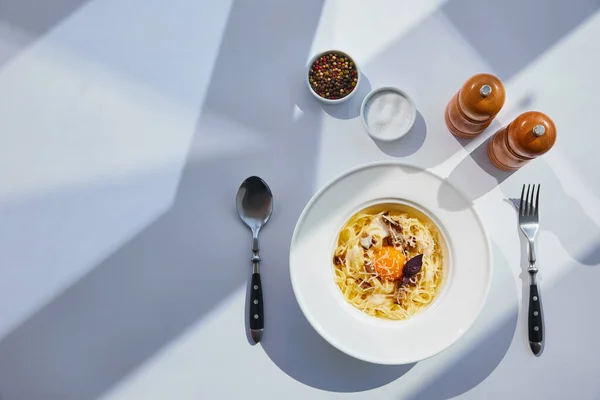 Vue de dessus de délicieuses pâtes carbonara servies avec couverts, moulins à sel et poivre sur table blanche au soleil — Photo de stock