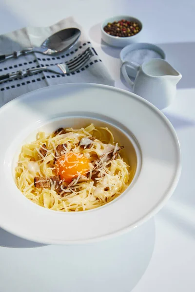 Deliziosa pasta alla carbonara servita con posate, panna e condimento su tavolo bianco alla luce del sole — Foto stock