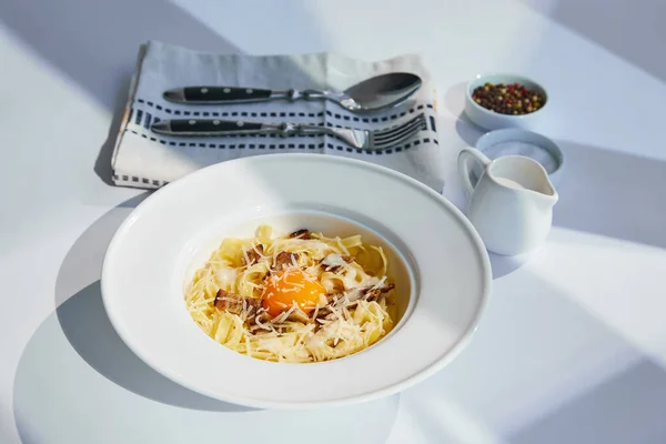 Deliziosa pasta alla carbonara servita con posate, panna e condimento su tavolo bianco alla luce del sole — Foto stock