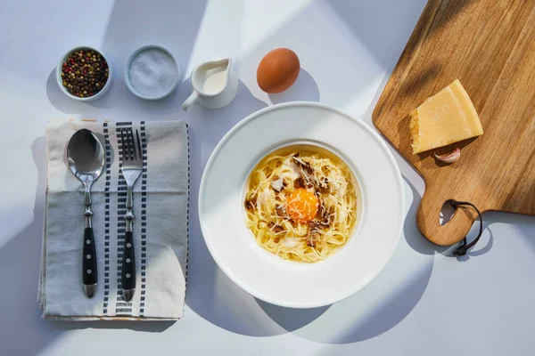 Vista superior de la deliciosa pasta carbonara servida con cubiertos, condimentos e ingredientes en la mesa blanca a la luz del sol - foto de stock