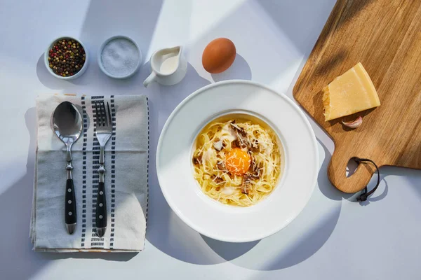 Верхний вид вкусной пасты карбонара подается со столовыми приборами, приправы и ингредиенты на белый стол в солнечном свете — стоковое фото