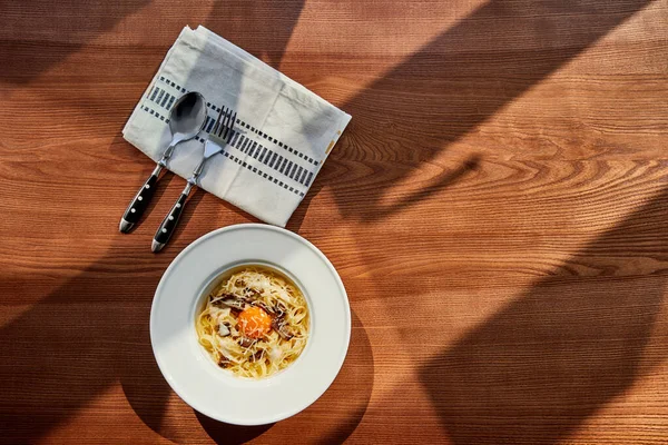 Blick von oben auf köstliche Pasta Carbonara serviert mit Besteck und Serviette auf Holztisch im Sonnenlicht — Stockfoto