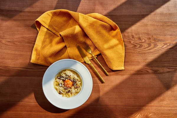 Vista superior de la deliciosa pasta carbonara servida con cubiertos dorados y servilleta amarilla en mesa de madera a la luz del sol - foto de stock