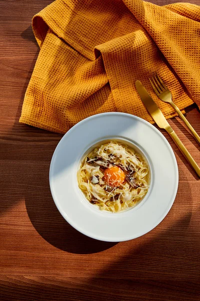 Blick von oben auf köstliche Pasta Carbonara serviert mit goldenem Besteck und gelber Serviette auf Holztisch im Sonnenlicht — Stockfoto