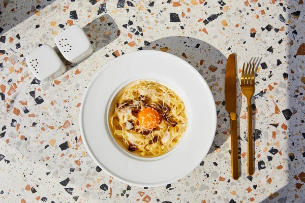 Vue de dessus de délicieuses pâtes carbonara servies avec des couverts dorés, salière et poivrière sur table en pierre au soleil — Photo de stock