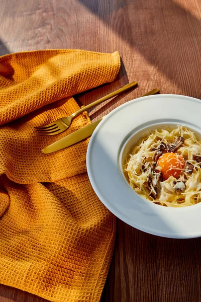 Délicieuses pâtes carbonara servi avec couverts dorés et serviette jaune sur table en bois à la lumière du soleil — Photo de stock