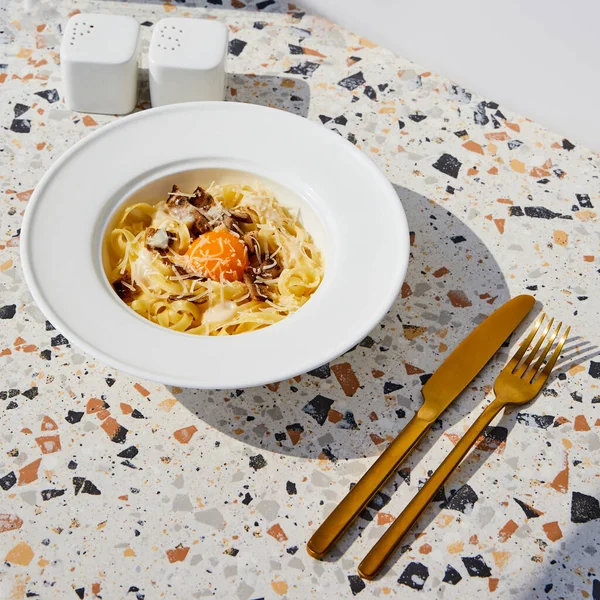 Délicieuses pâtes carbonara servies avec couverts dorés, salière et poivrière sur table en pierre au soleil — Photo de stock