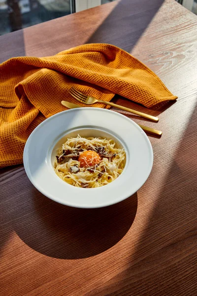 Délicieuses pâtes carbonara servi avec couverts dorés et serviette jaune sur table en bois à la lumière du soleil — Stock Photo