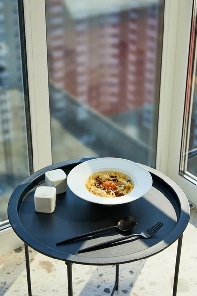 Délicieuses pâtes carbonara servies avec des couverts, assaisonnement sur table noire sur fond de marbre à la lumière du soleil près de la fenêtre — Photo de stock