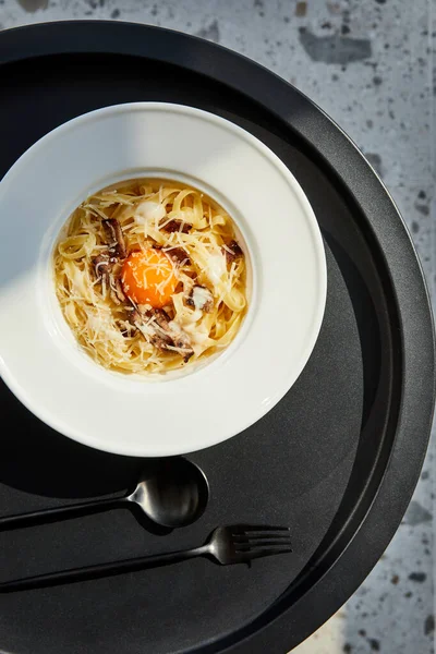 Top view of delicious pasta carbonara served with cutlery on black table on marble background in sunlight — Stock Photo