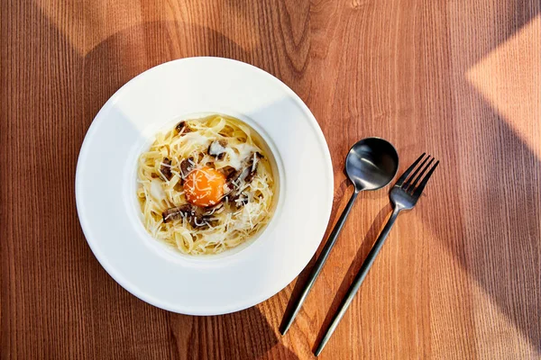 Vista superior de la deliciosa pasta carbonara servida con cubiertos negros sobre fondo de madera a la luz del sol - foto de stock