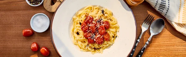 Top view of delicious pasta with tomatoes served on wooden table with cutlery, napkin, seasoning and napkin in sunlight, panoramic shot — Stock Photo