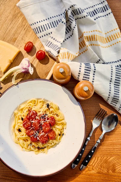 Vista dall'alto di deliziose paste con pomodori servite su tavolo in legno con posate, tovagliolo, condimento e ingredienti alla luce del sole — Foto stock