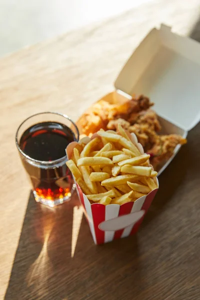 Fuoco selettivo di pollo fritto in profondità, patatine fritte e soda in vetro sul tavolo in legno alla luce del sole — Foto stock