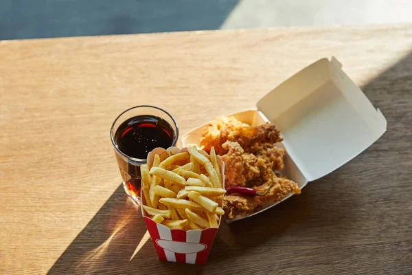 Pollo fritto in olio, patatine fritte e soda in vetro sul tavolo di legno alla luce del sole — Foto stock