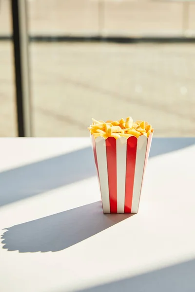 Tasty french fries in striped bucket on white table in sunlight near window — Stock Photo