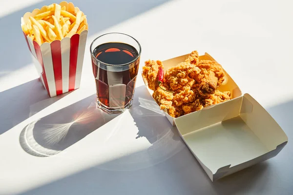 Gustoso pollo fritto, patatine fritte e soda in vetro su tavolo bianco alla luce del sole — Foto stock