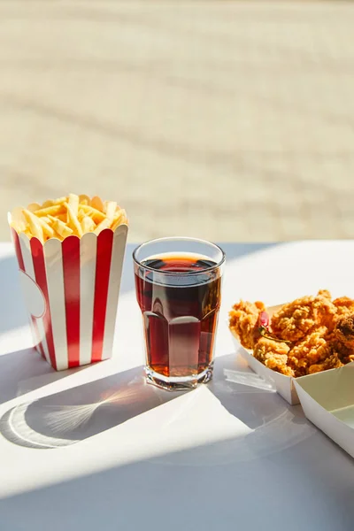 Sabroso pollo frito, papas fritas y refrescos en vidrio sobre mesa blanca a la luz del sol cerca de la ventana - foto de stock