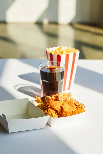 Selektiver Fokus von leckerem frittiertem Hähnchen, Pommes frites und Limo im Glas auf weißem Tisch im Sonnenlicht — Stockfoto