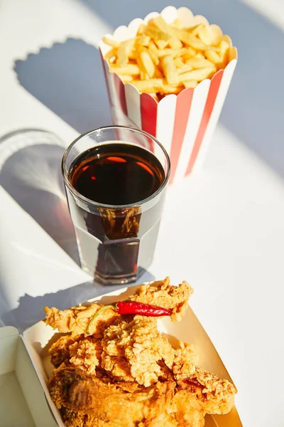 Selektiver Fokus von leckerem frittiertem Hähnchen, Pommes frites und Limo im Glas auf weißem Tisch im Sonnenlicht — Stockfoto