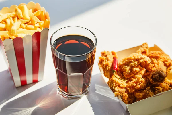 Gustoso pollo fritto, patatine fritte e soda in vetro su tavolo bianco alla luce del sole — Foto stock