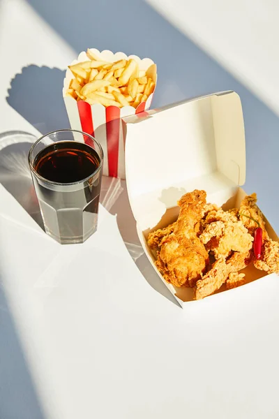 Tasty deep fried chicken, french fries and soda in glass on white table in sunlight — Stock Photo