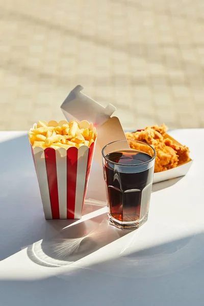 Enfoque selectivo de sabroso pollo frito, papas fritas y refrescos en vidrio sobre mesa blanca a la luz del sol cerca de la ventana - foto de stock