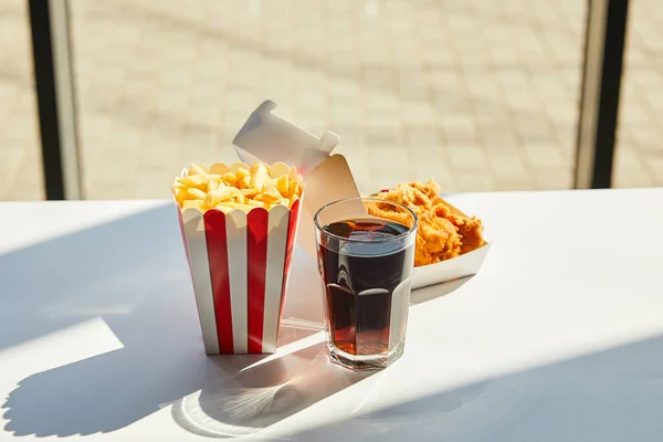 Tasty deep fried chicken, french fries and soda in glass on white table in sunlight near window — Stock Photo