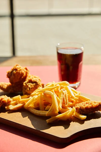 Selective focus of crispy deep fried chicken and french fries served on wooden cutting board with soda in sunlight near window — Stock Photo