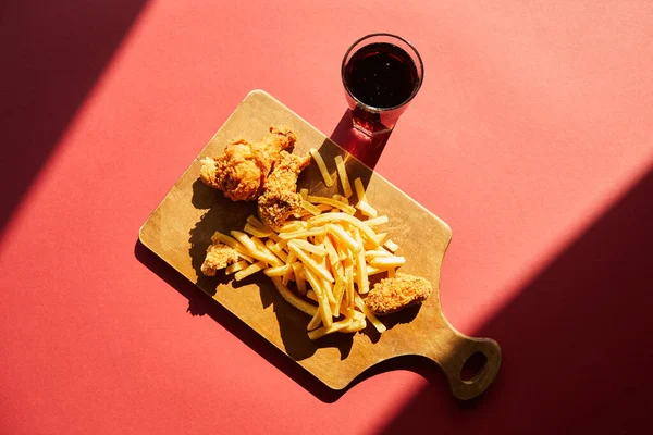 Top view of crispy deep fried chicken and french fries served on wooden cutting board with soda in sunlight — Stock Photo