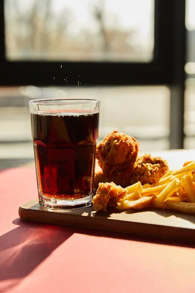 Crispy deep fried chicken and french fries served on wooden cutting board with soda in sunlight near window — Stock Photo