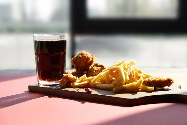 Pollo fritto croccante e patatine fritte servite sul tagliere di legno con soda alla luce del sole vicino alla finestra — Foto stock