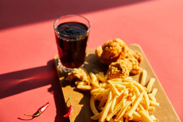 Selective focus of spicy deep fried chicken and french fries served on wooden cutting board with soda in sunlight — Stock Photo