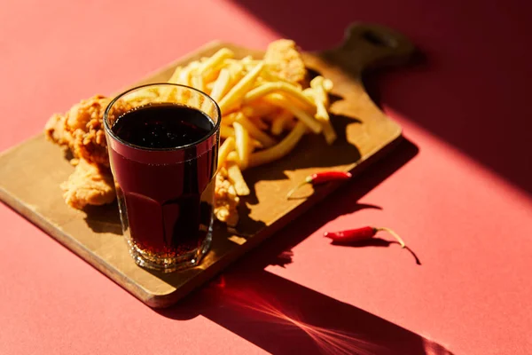 Enfoque selectivo de pollo frito picante y papas fritas servidas en tabla de cortar de madera con soda a la luz del sol - foto de stock