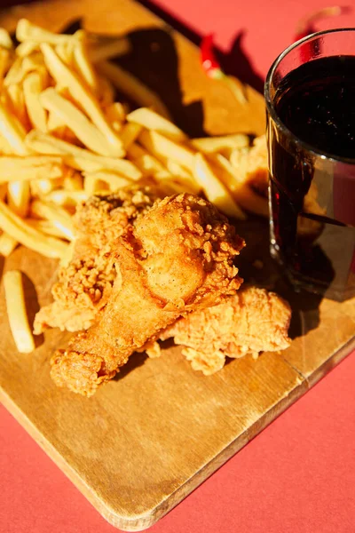 Foco seletivo de frango frito crocante e batatas fritas servidas em tábua de corte de madeira com refrigerante à luz solar — Fotografia de Stock