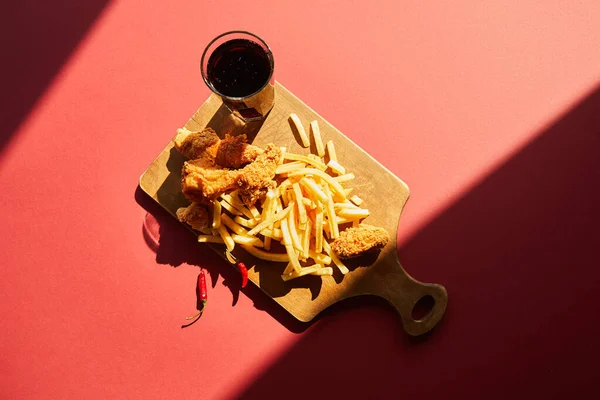 Top view of spicy deep fried chicken and french fries served on wooden cutting board with soda in sunlight — Stock Photo