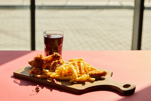 Spicy deep fried chicken and french fries served on wooden cutting board with soda in sunlight near window — Stock Photo