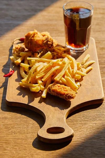 Selective focus of spicy deep fried chicken, french fries on board with soda in glass on wooden table in sunlight — Stock Photo