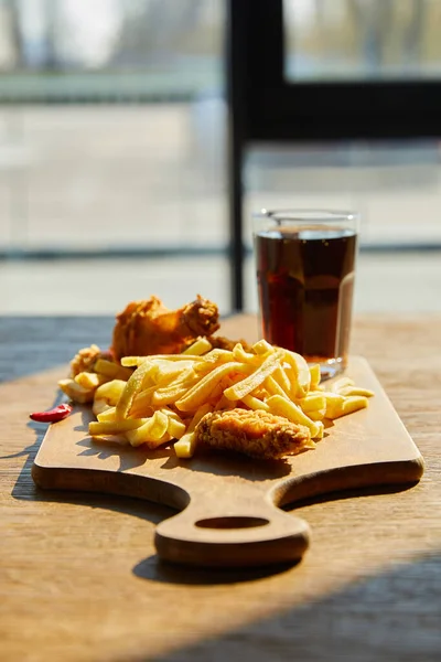 Fuoco selettivo di pollo fritto in profondità piccante, patatine fritte a bordo con soda in vetro su tavolo di legno alla luce del sole vicino alla finestra — Foto stock