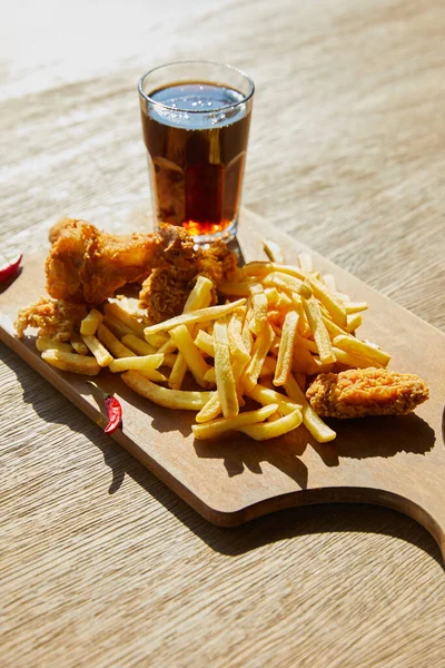 Spicy deep fried chicken, french fries on board with soda in glass on wooden table in sunlight — Stock Photo