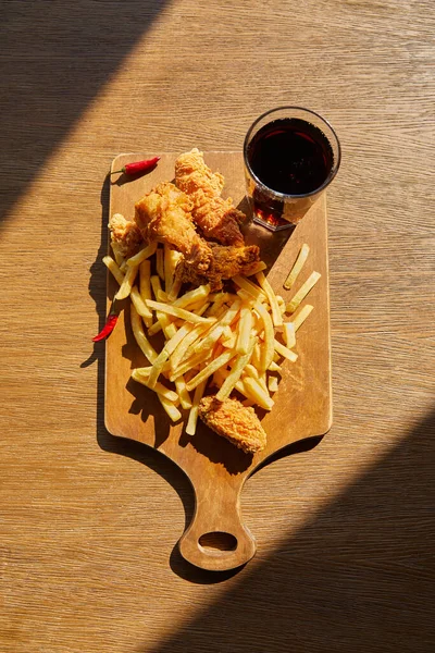 Top view of spicy deep fried chicken, french fries on board with soda in glass on wooden table in sunlight — Stock Photo