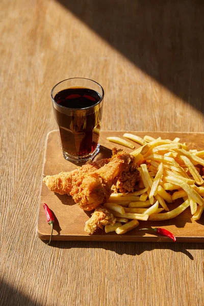 Spicy deep fried chicken, french fries on board with soda in glass on wooden table in sunlight — Stock Photo